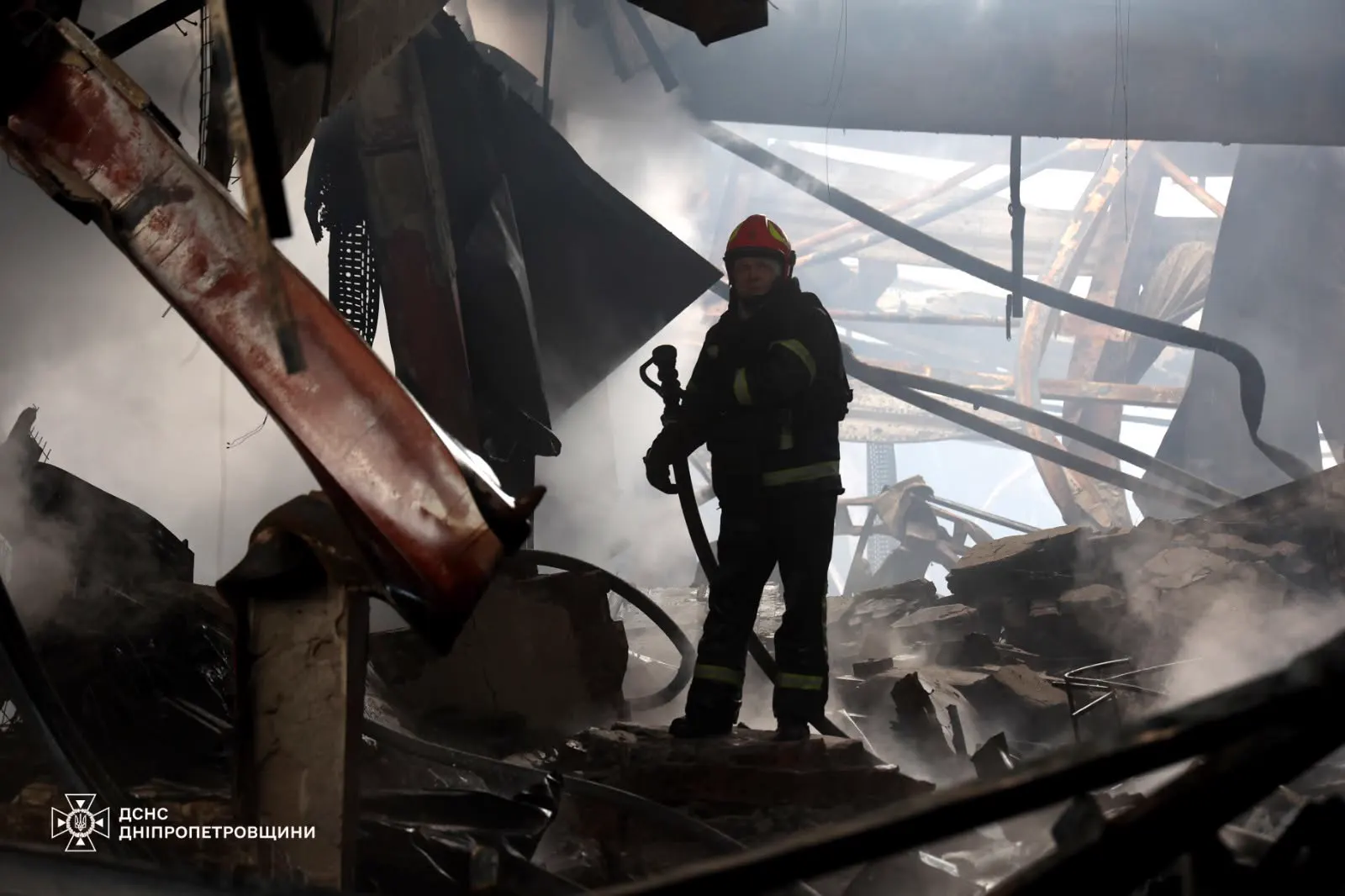 Un bombero con casco rojo sostiene una manguera de pie sobre los escombros humeantes de una nave incendiada, entre vigas retorcidas y planchas metálicas caídas.