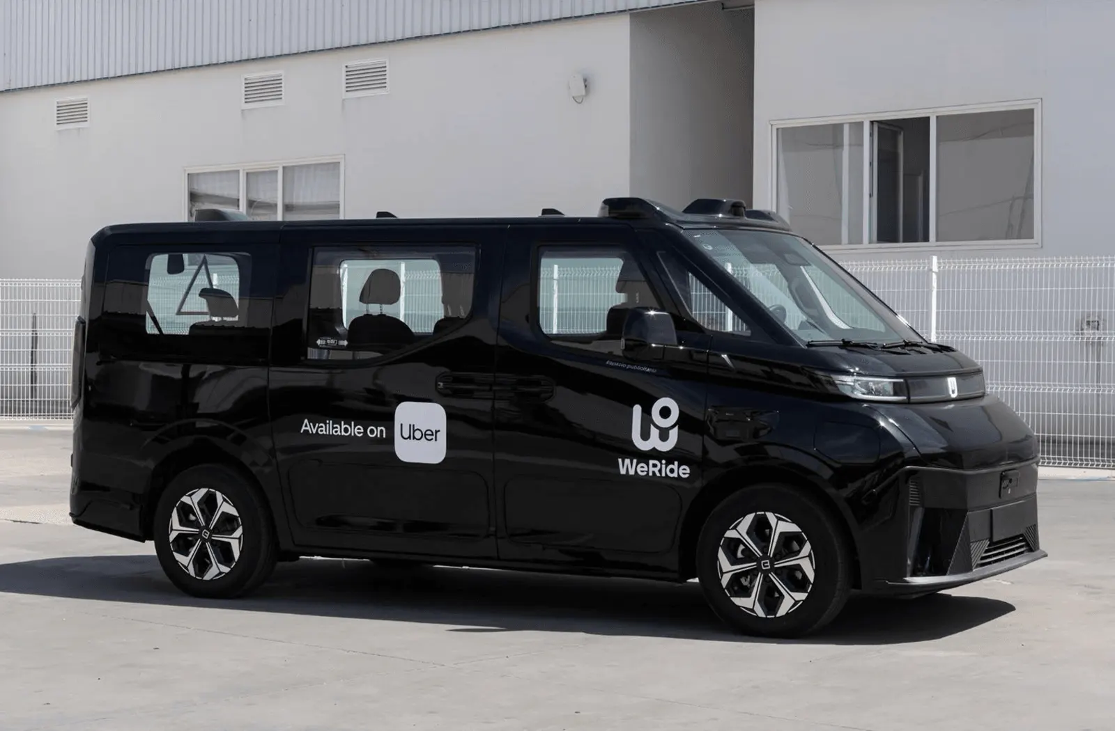 Monovolumen negro de WeRide con sensores en el techo y el rótulo «Available on Uber» en la puerta lateral, aparcado junto a una nave.