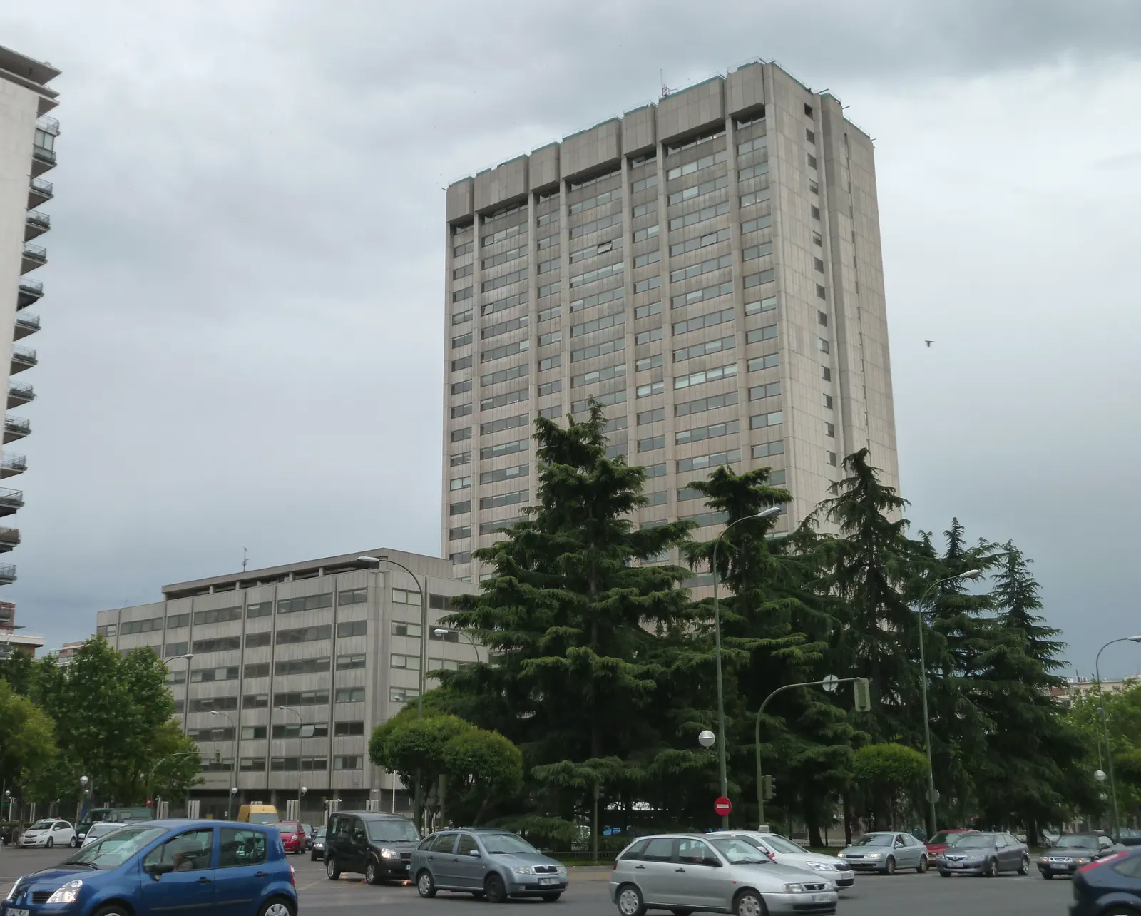 Torre de oficinas de hormigón de más de veinte plantas bajo un cielo encapotado, con un bloque bajo de ventanas corridas a su izquierda, cedros en primer término y tráfico de coches en la avenida que pasa por delante.