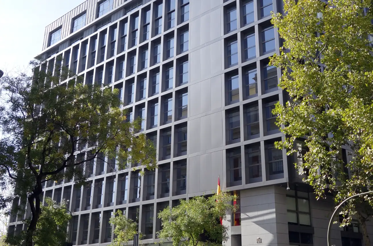 Fachada de un edificio moderno de oficinas de varias plantas, con ventanas cuadradas enmarcadas en piedra clara y paneles metálicos, vista a contrapicado entre las copas de dos árboles; en la esquina inferior derecha ondea una bandera de España junto a un escudo grabado en el muro.