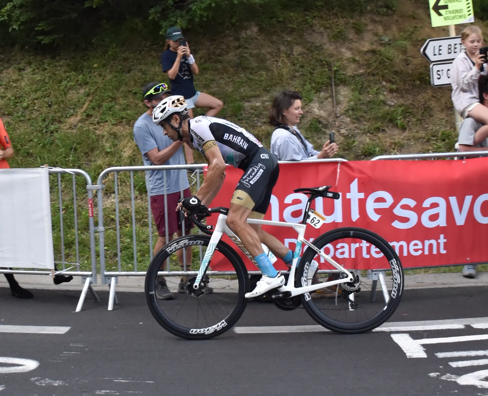 Un ciclista con maillot blanco y negro del Bahrain Victorious y dorsal 62 pedalea de pie sobre los pedales por una carretera de montaña, entre vallas y espectadores que lo fotografían.