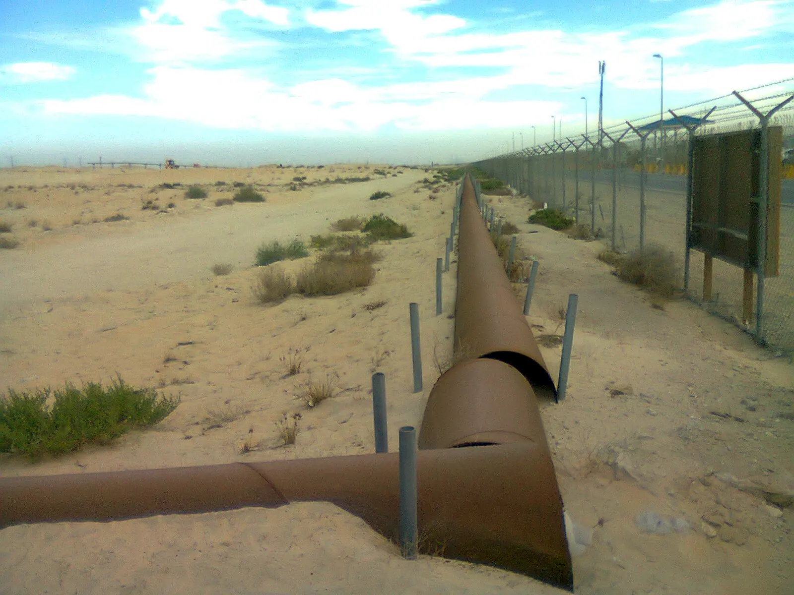 Dos tuberías de acero oxidado recorren un terreno arenoso junto a una valla metálica rematada con alambre de espino, bajo un cielo con nubes, en el desierto de Jubail (Arabia Saudí).