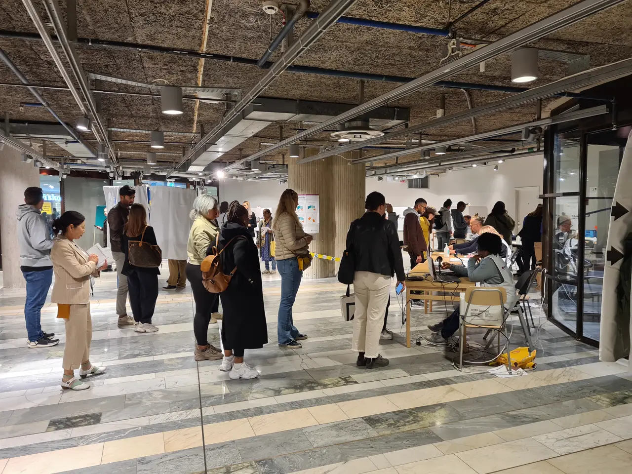 Una veintena de personas hacen cola en el vestíbulo de un edificio público sueco habilitado como local de voto anticipado; al fondo, a la derecha, funcionarios electorales atienden en mesas con ordenadores junto a varias cabinas de votación blancas.