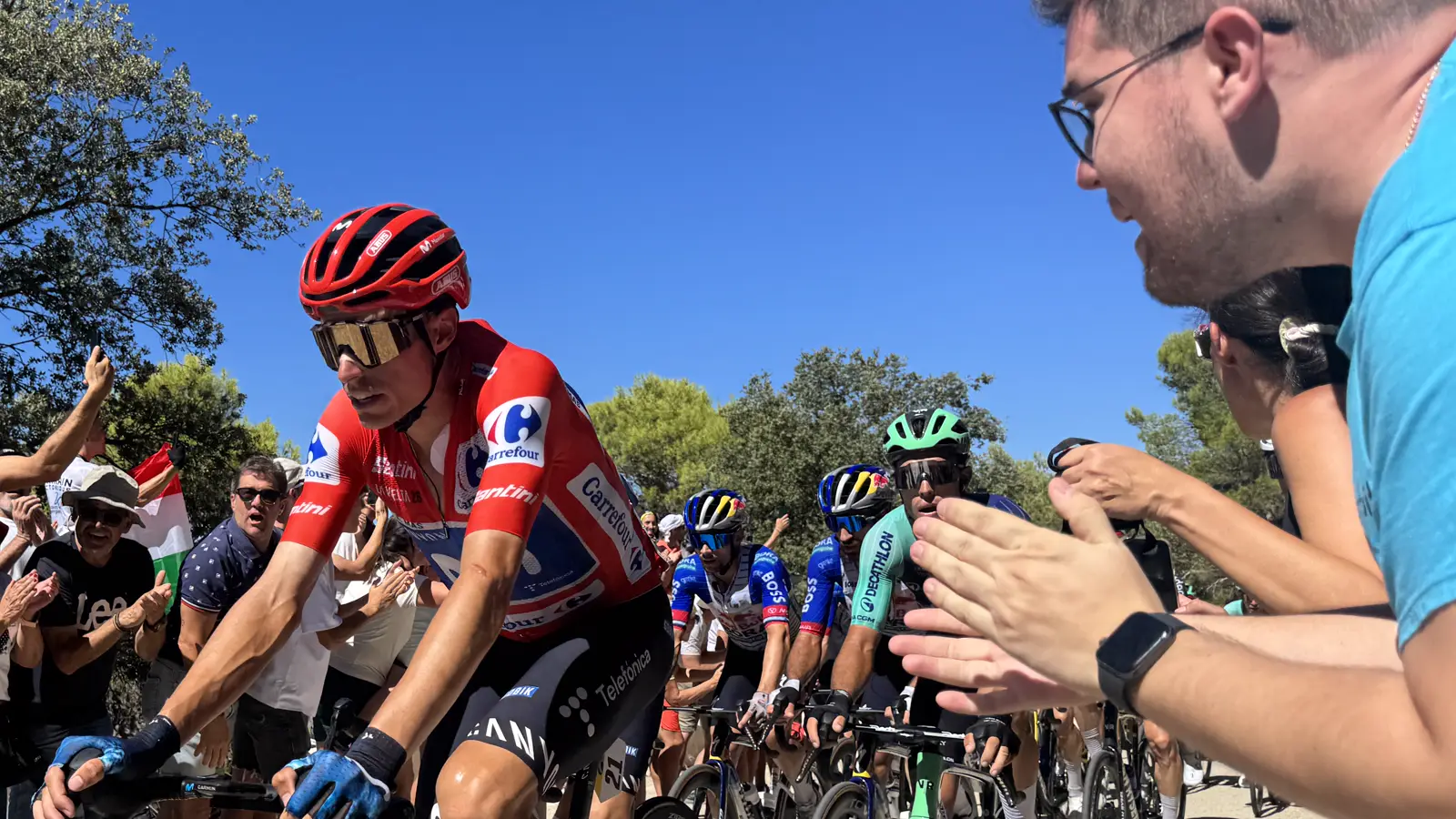 Un ciclista con el maillot rojo de líder de la Vuelta, casco rojo y gafas doradas sube una carretera de montaña de pie sobre los pedales, rodeado de espectadores que aplauden; detrás le siguen otros corredores en fila.