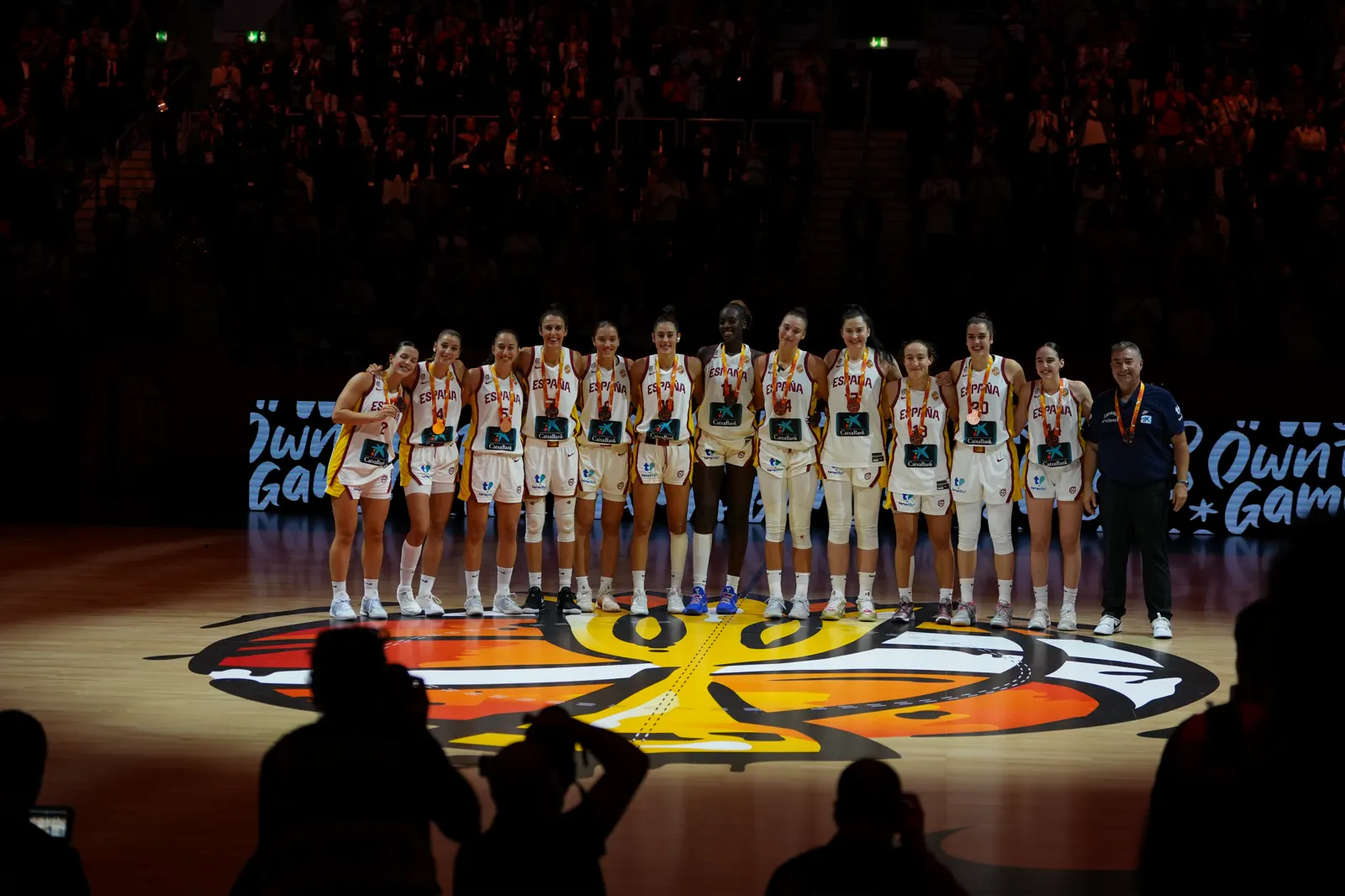 Las doce jugadoras de la selección española, con camiseta blanca y la medalla de bronce al cuello, posan en fila sobre el círculo central de la pista junto al seleccionador; al fondo, las gradas llenas en penumbra