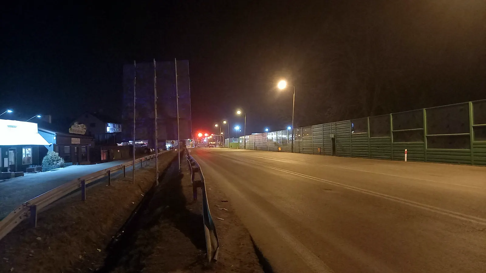 Carretera de acceso al paso fronterizo de Dorohusk-Jagodzin de noche: calzada vacía bajo farolas anaranjadas, un semáforo en rojo al fondo, vallas metálicas verdes a la derecha y casas bajas iluminadas a la izquierda.