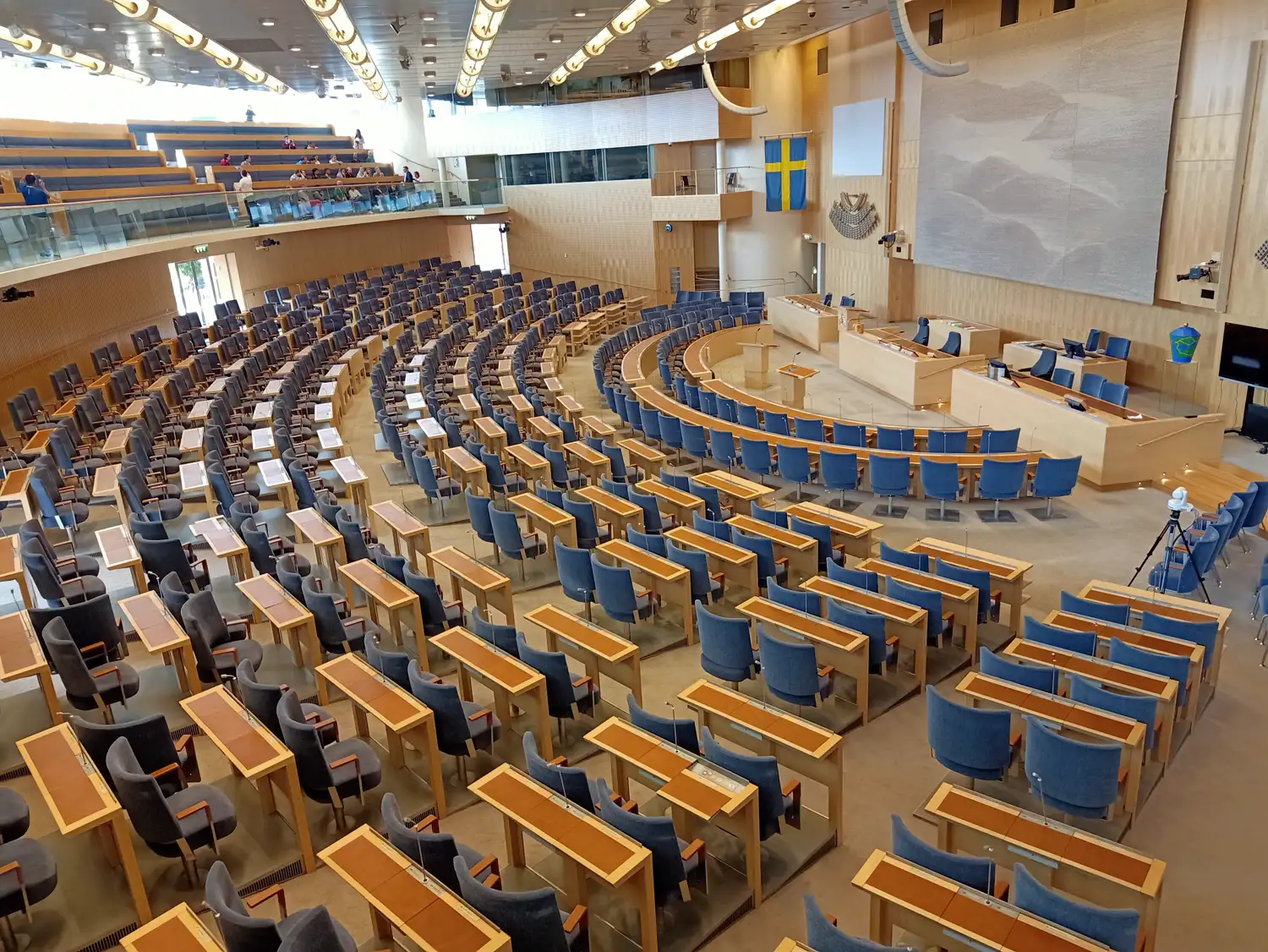 Vista elevada del hemiciclo vacío del Riksdag sueco: filas curvas de pupitres de madera clara con sillones azules, la mesa de la presidencia al fondo y una bandera de Suecia colgada en la pared.