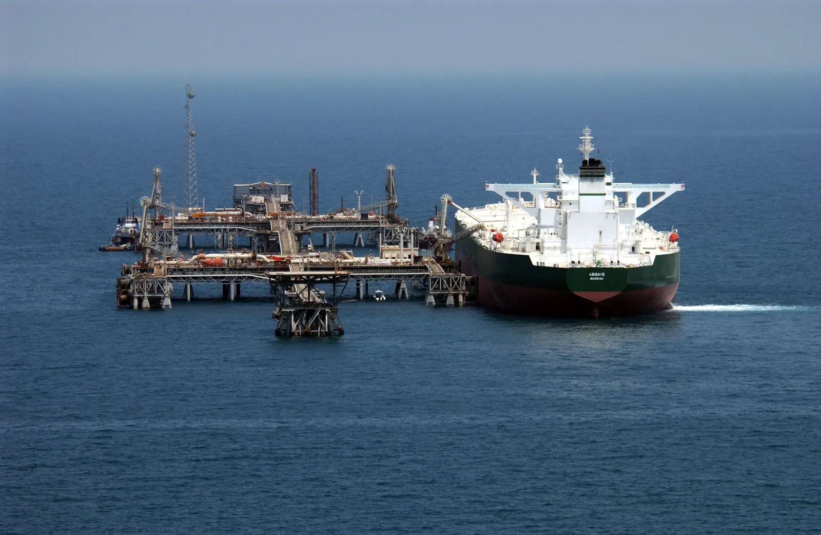 Vista aérea de un petrolero de casco verde y rojo, con el nombre «Abqaiq» en la proa, amarrado a una terminal petrolera de estructuras metálicas montada sobre pilotes en mitad del mar.