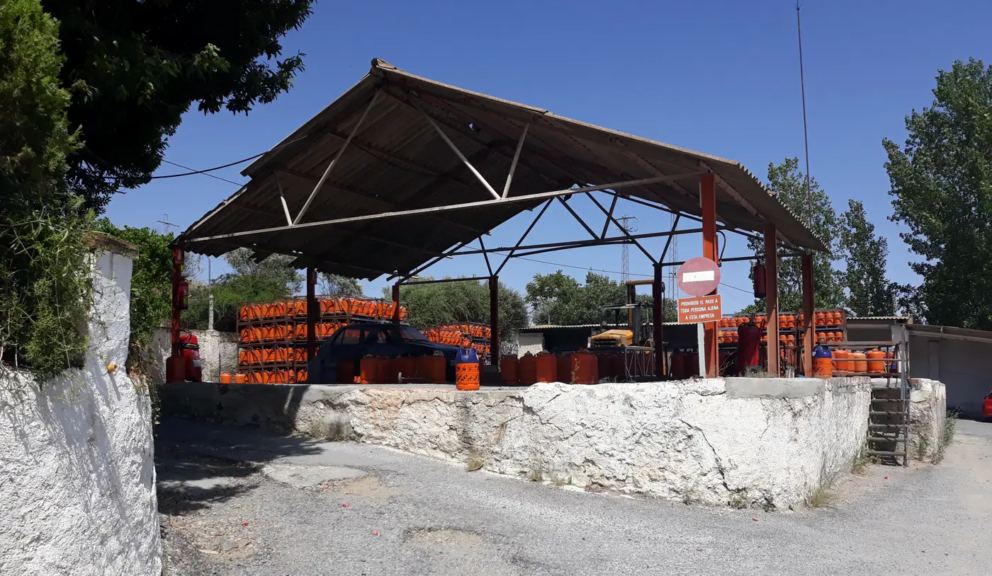 Depósito de bombonas de butano al aire libre bajo una nave de chapa sostenida por vigas metálicas: centenares de bombonas naranjas apiladas en jaulas, un coche azul aparcado dentro del recinto, un cartel rojo de prohibido el paso y un muro encalado en primer término, bajo un cielo azul sin nubes.