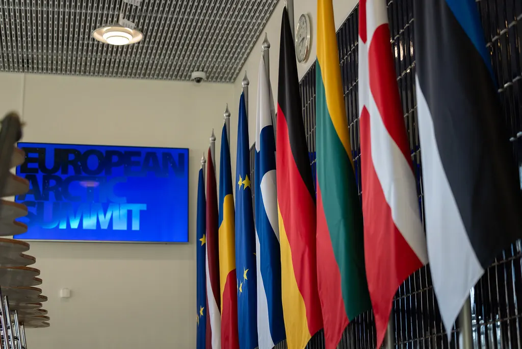Banderas nacionales y de la Unión Europea alineadas junto a una pantalla azul con el rótulo «European Arctic Summit» en la sala de la cumbre