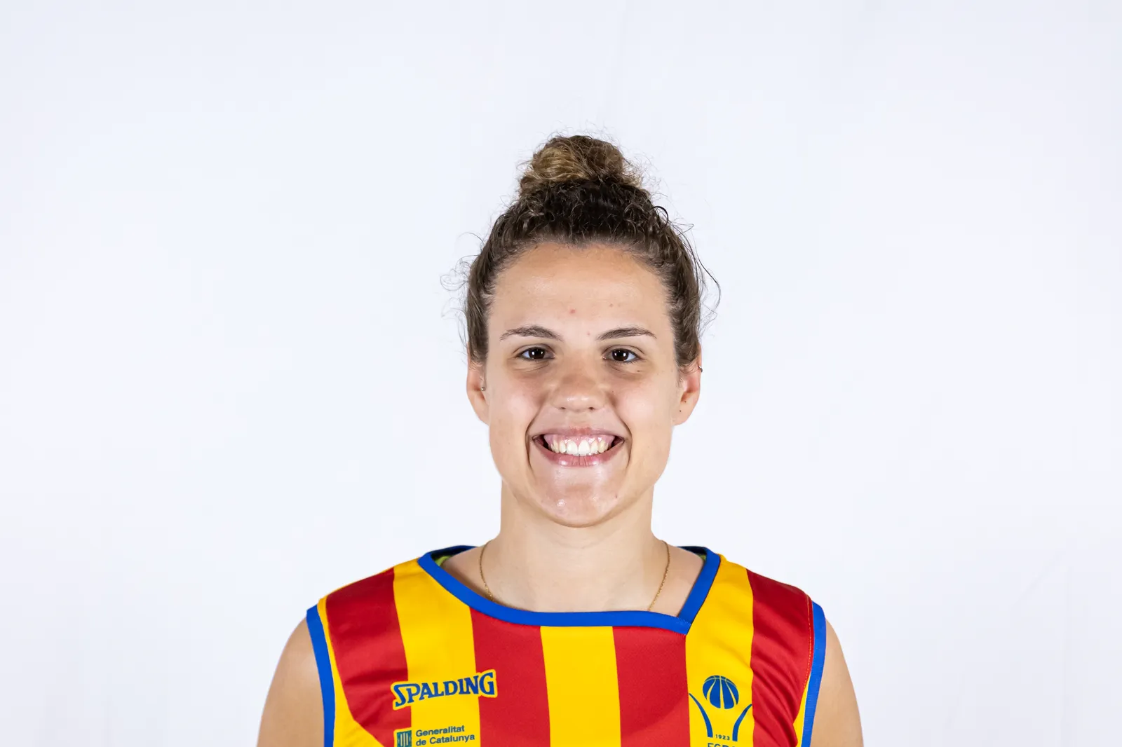 Retrato de la jugadora de baloncesto Txell Alarcón, sonriente, con una camiseta de tirantes a franjas rojas y amarillas sobre fondo blanco