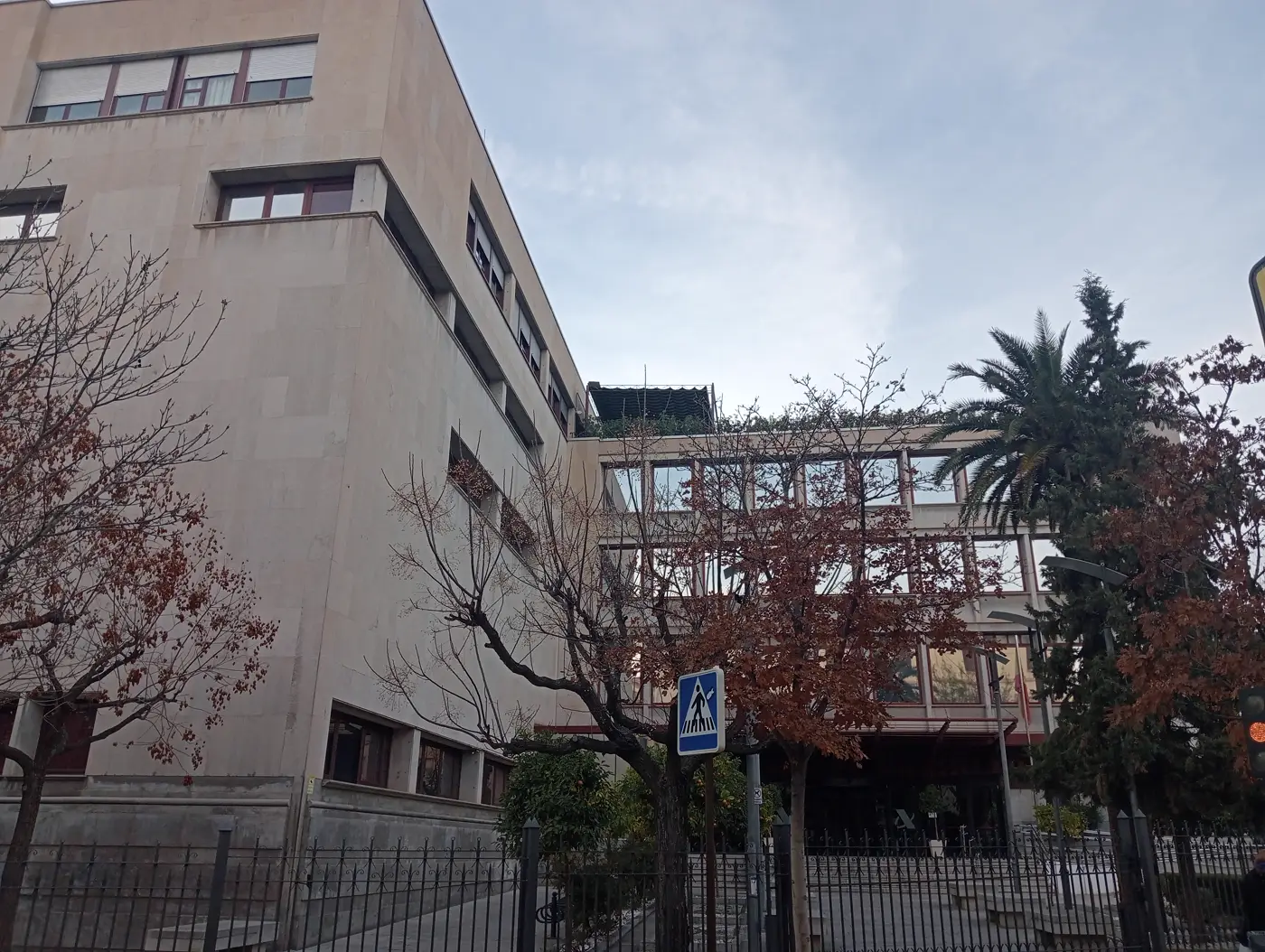 Fachada de piedra clara de un edificio administrativo de varias plantas, con ventanas de marco granate y un cuerpo acristalado a la derecha. Delante, árboles sin hoja, una palmera, una verja metálica sobre un murete y una señal azul de paso de peatones; el cielo está nublado.