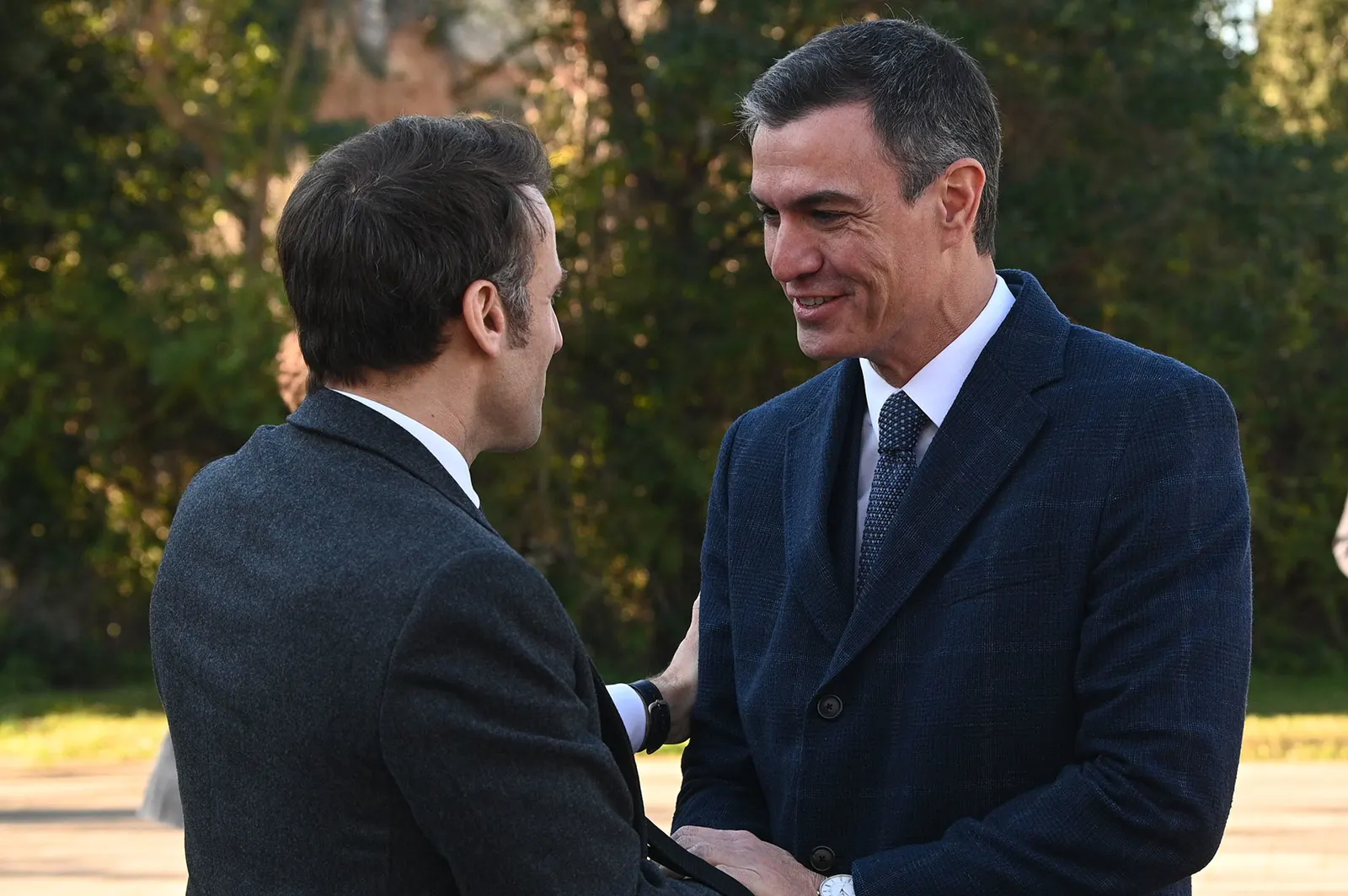 Pedro Sánchez, sonriente y de frente, saluda con la mano en el hombro a Emmanuel Macron, de espaldas y de perfil, ambos con abrigo oscuro y ante una zona ajardinada.