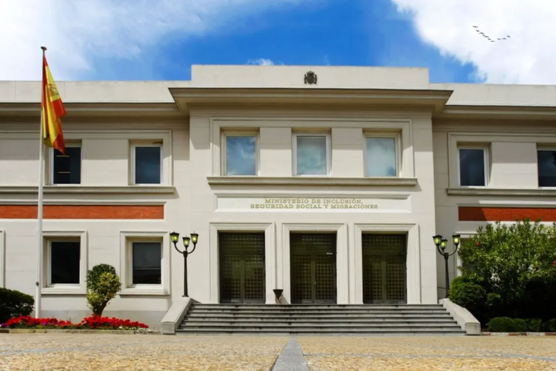 Fachada blanca de dos plantas del Ministerio de Inclusión, Seguridad Social y Migraciones, con la bandera de España a la izquierda, escalinata de acceso y el nombre del ministerio grabado en letras doradas sobre la entrada