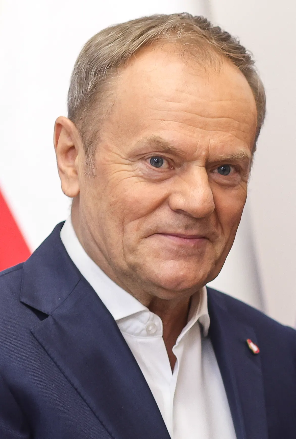Retrato de Donald Tusk, de pelo corto y canoso, con americana azul marino y camisa blanca sin corbata, con un pin de la bandera polaca en la solapa y una bandera blanquirroja desenfocada al fondo.