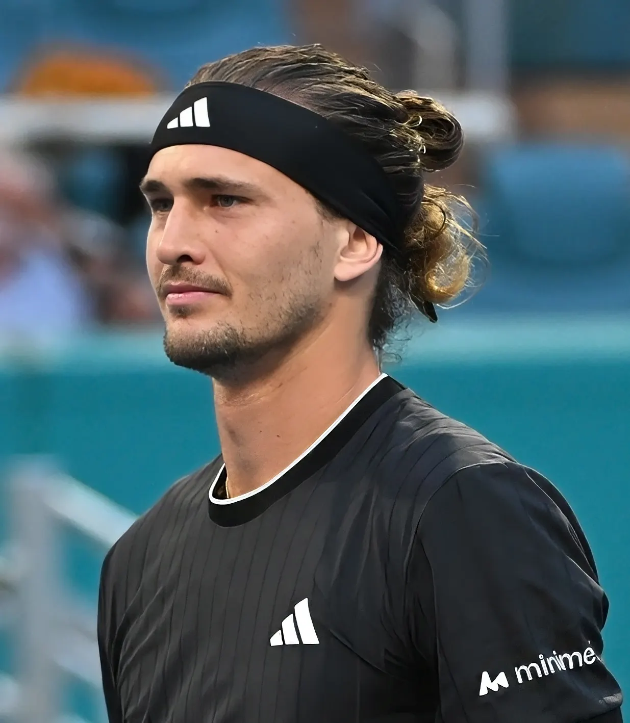 Primer plano de un tenista con cinta negra en la cabeza, el pelo recogido en un moño y camiseta negra, mirando al frente en una pista de tenis con gradas azules desenfocadas al fondo.