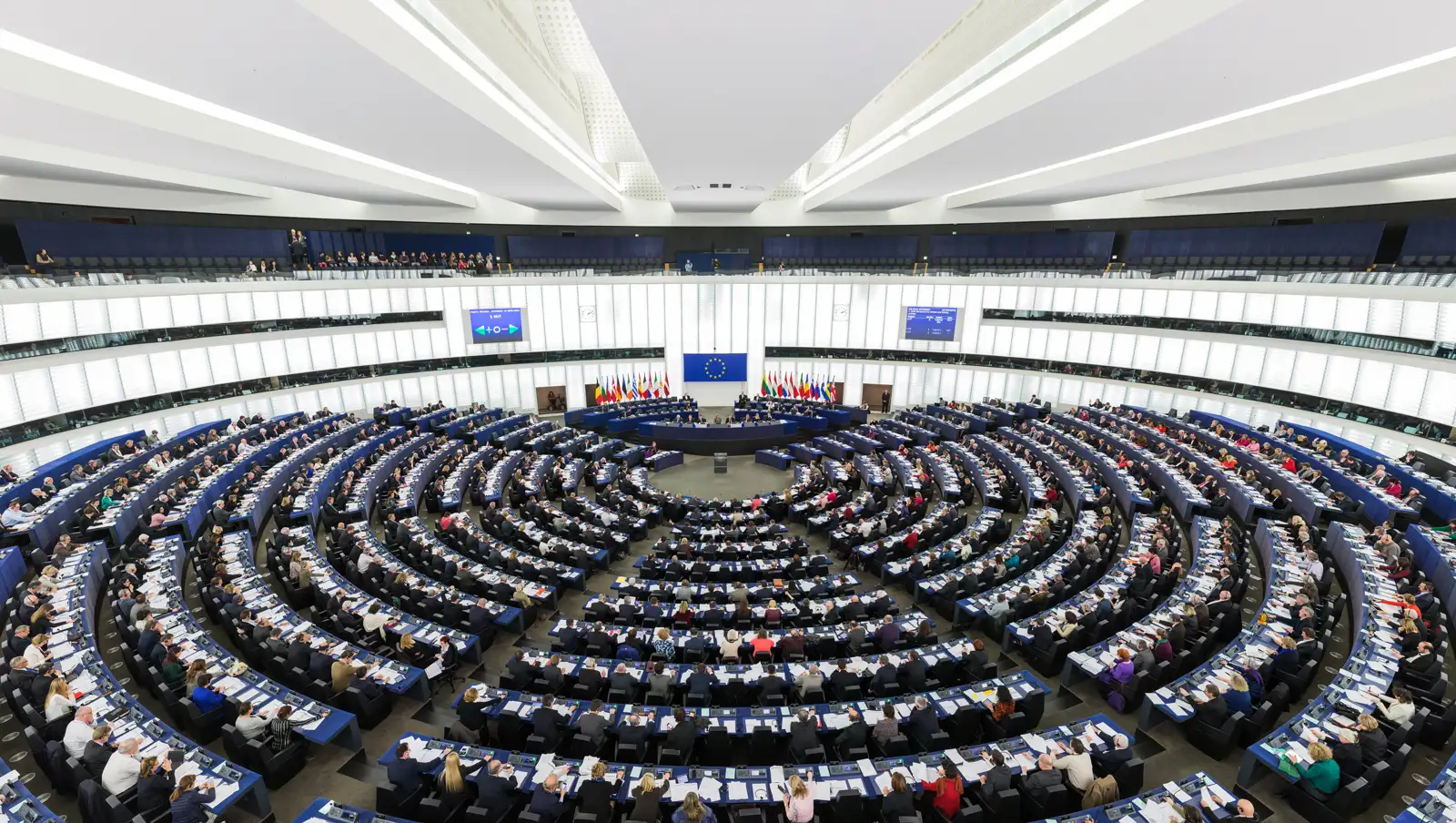 Vista general del hemiciclo del Parlamento Europeo en Estrasburgo durante una sesión plenaria: filas curvas de escaños azules ocupadas por cientos de eurodiputados y, al fondo, la mesa de la presidencia bajo la bandera de la Unión Europea y las de los Estados miembros.